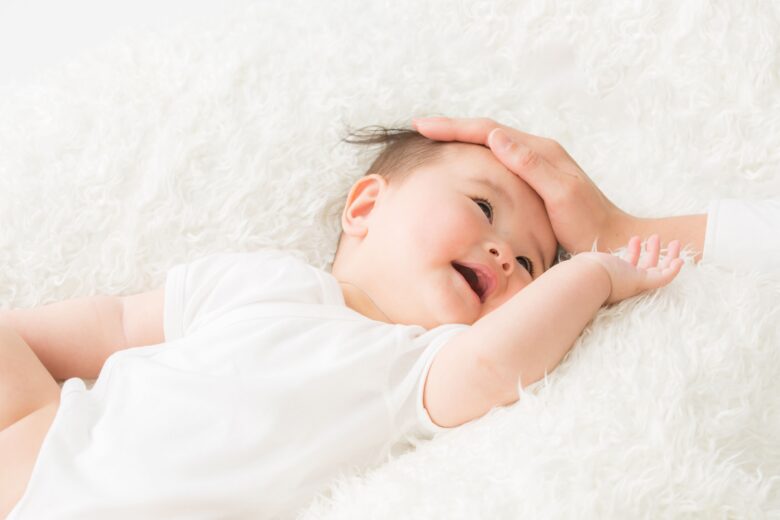柔らかな布の上にくつろぐ赤ちゃんと、赤ちゃんのおでこに手を添える親子の様子